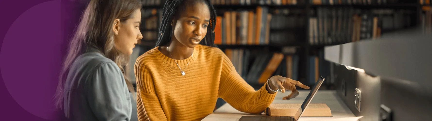 Two young women at a desk in a library, discussing what they see on a Lenovo laptop